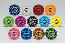 A set of circular gaming chips arranged in three rows on a light gray background. The chips come in multiple colors including blue, silver, red, orange, green, pink, black, light blue, yellow, brown, and dark green, each with different denominations from 25 to 100,000.